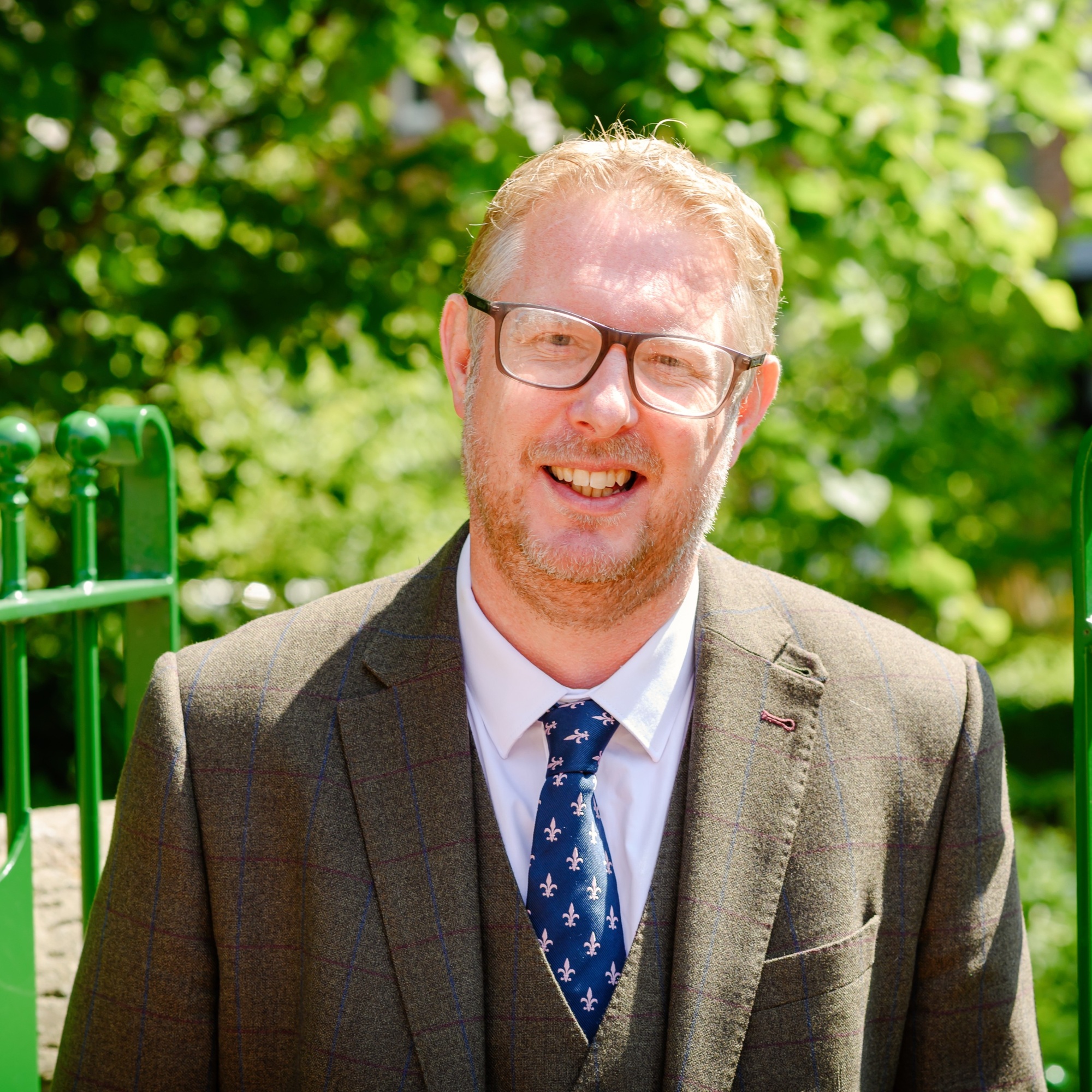 Photograph of John Sample in front of school gates. He wears a brown suit and is smiling.